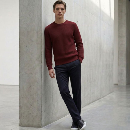 Man wearing a red sweater and dark pants standing against a concrete wall.
