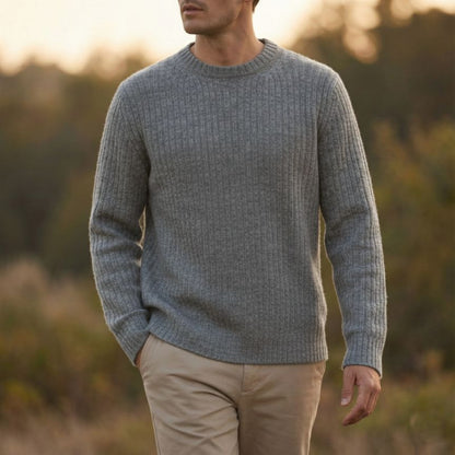 Man wearing a gray sweater and beige pants standing in a natural setting with blurred background